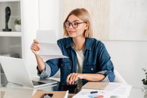 Young caucasian woman paying domestic bills, making calculations at home desk. Female holding billing overdue, counting funds online, loan and debt